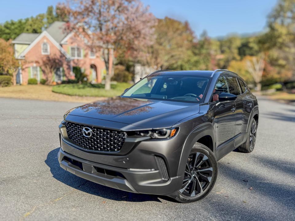 The left front of corner of a gray 2025 Mazda CX-50 Hybrid Premium Plus compact SUV parked in front of a house.