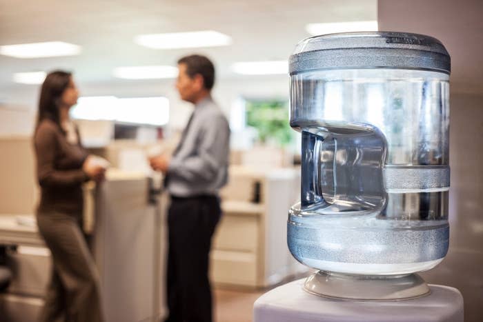 Office water cooler with two people in the background talking, suggesting a casual work setting or break-time conversation