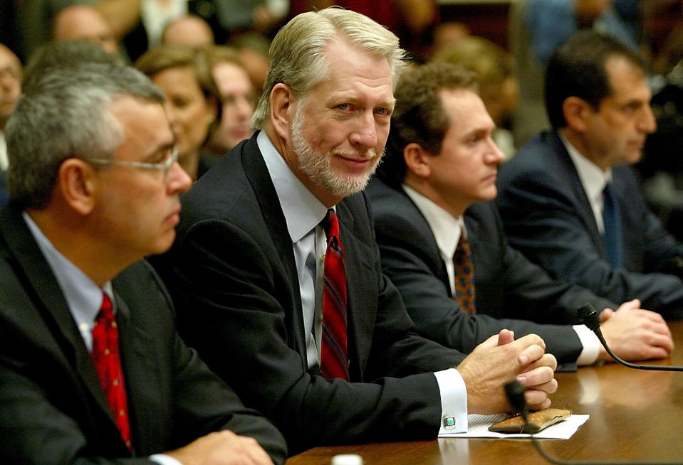 A group of men in suits sit at a conference table attentively listening, with one man looking directly at the camera