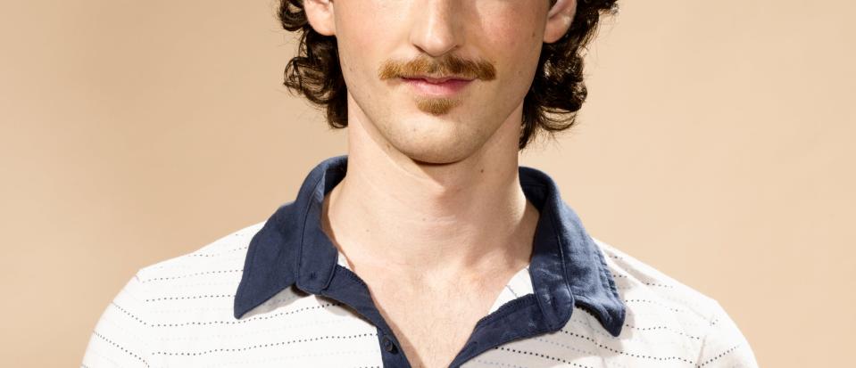 Person with curly hair and mustache, wearing a collared casual striped shirt, smiles at the camera. Plain background