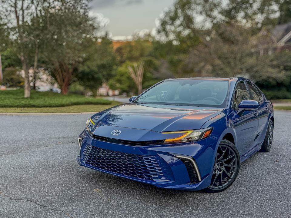 The left front corner of a blue 2025 Toyota Camry XSE AWD hybrid sedan parked in front of a row of homes.