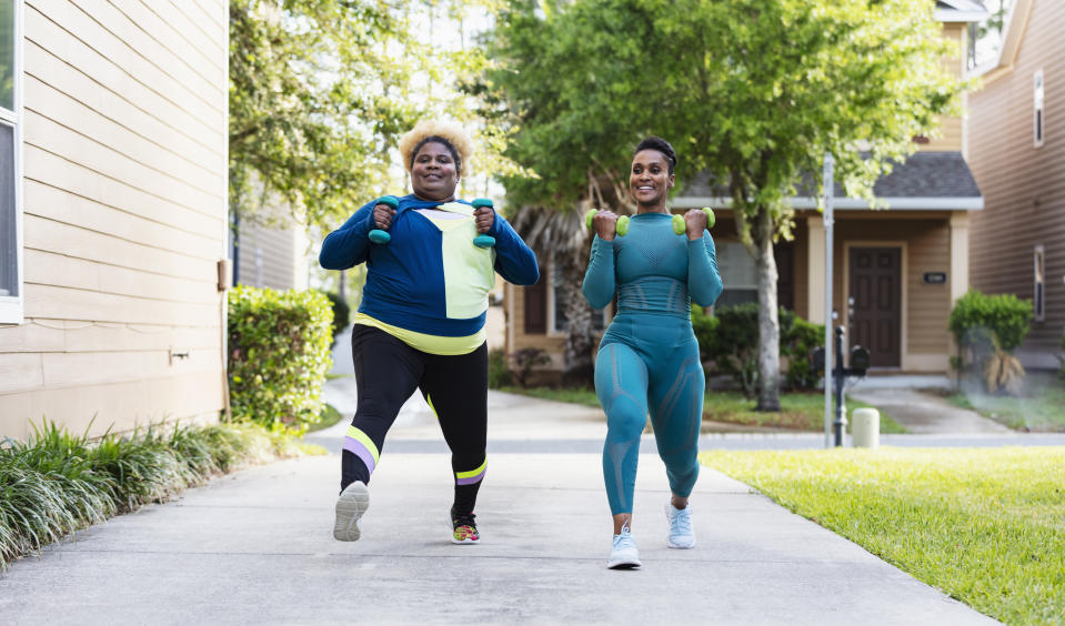 Two people walk outdoors in athletic wear, each holding dumbbells, smiling as they exercise in a suburban neighborhood