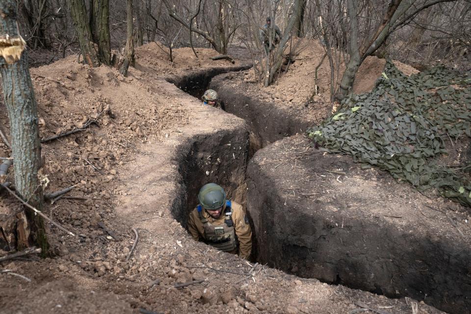 Two Ukrainian troops in a trench surrounded by brown earth and damaged trees.