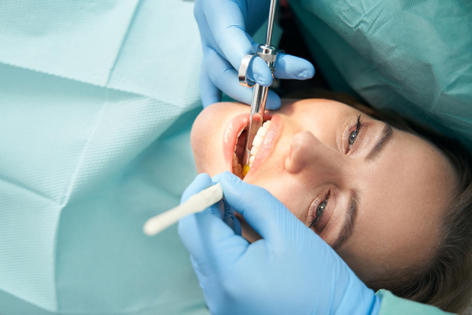 Person receiving dental care, lying back with a dental tool in their mouth, attended by a professional wearing gloves