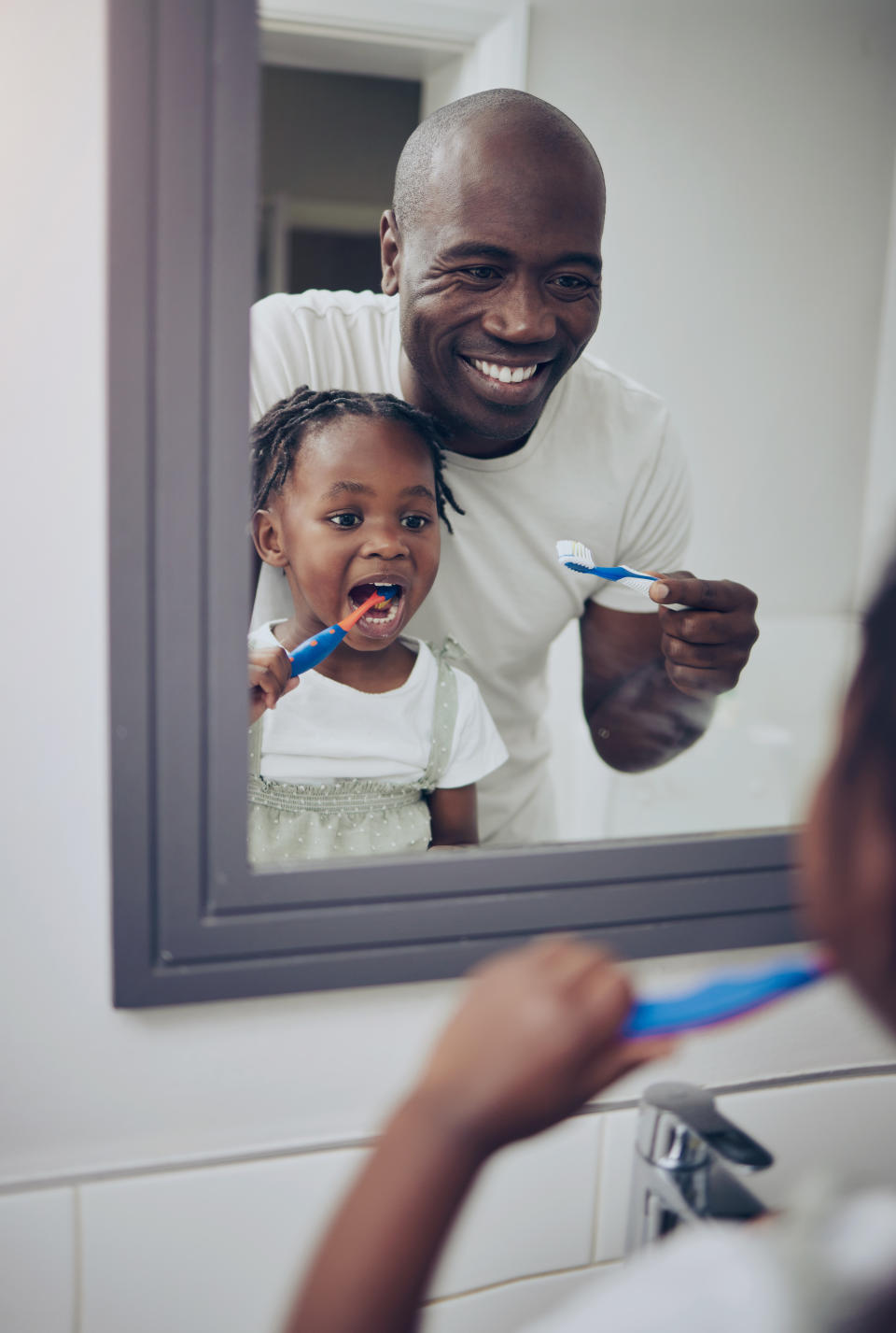 Father and daughter happily brush their teeth together in front of a bathroom mirror