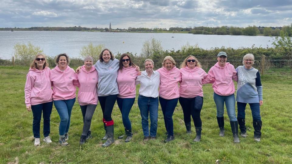 A row of woman &#x002013; some wearing pink hoodies &#x002013; standing on grass while smiling at the camera