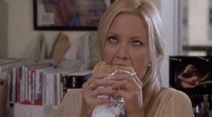 Person in an office setting eating a sandwich with an exasperated expression, surrounded by books and a poster in the background