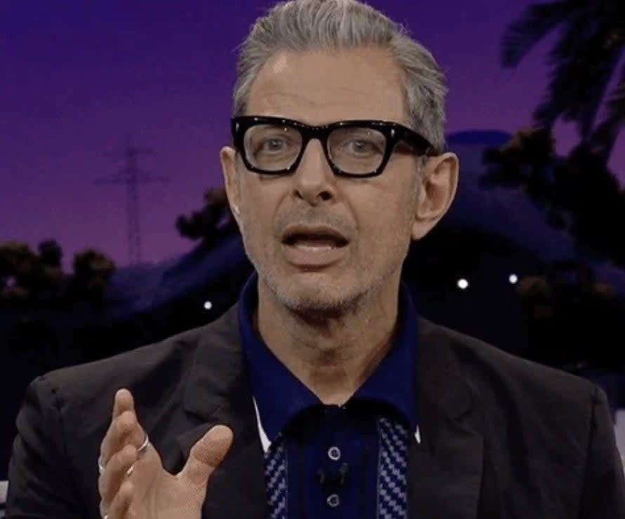 Person speaking, wearing glasses and a collared shirt under a suit jacket, with a blurred nighttime background