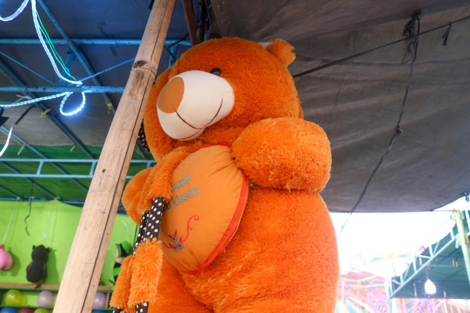 Large teddy bear holding a heart-shaped cushion with text, displayed at an amusement stall with game prizes visible in the background