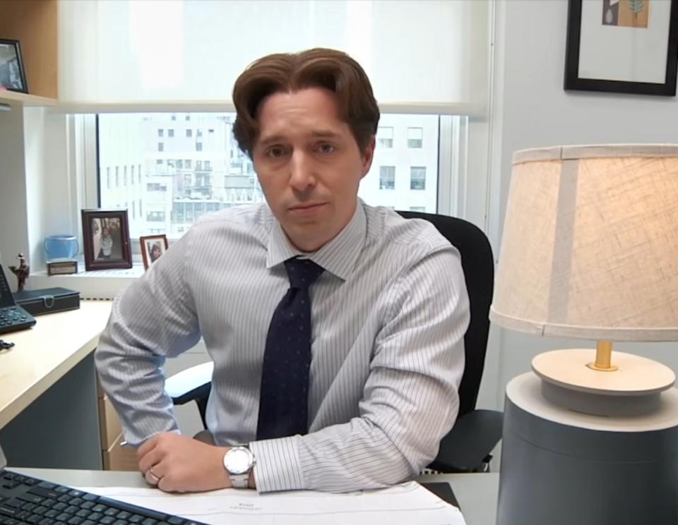 A man sits in an office at a desk with papers, a computer, and a lamp, looking directly at the camera