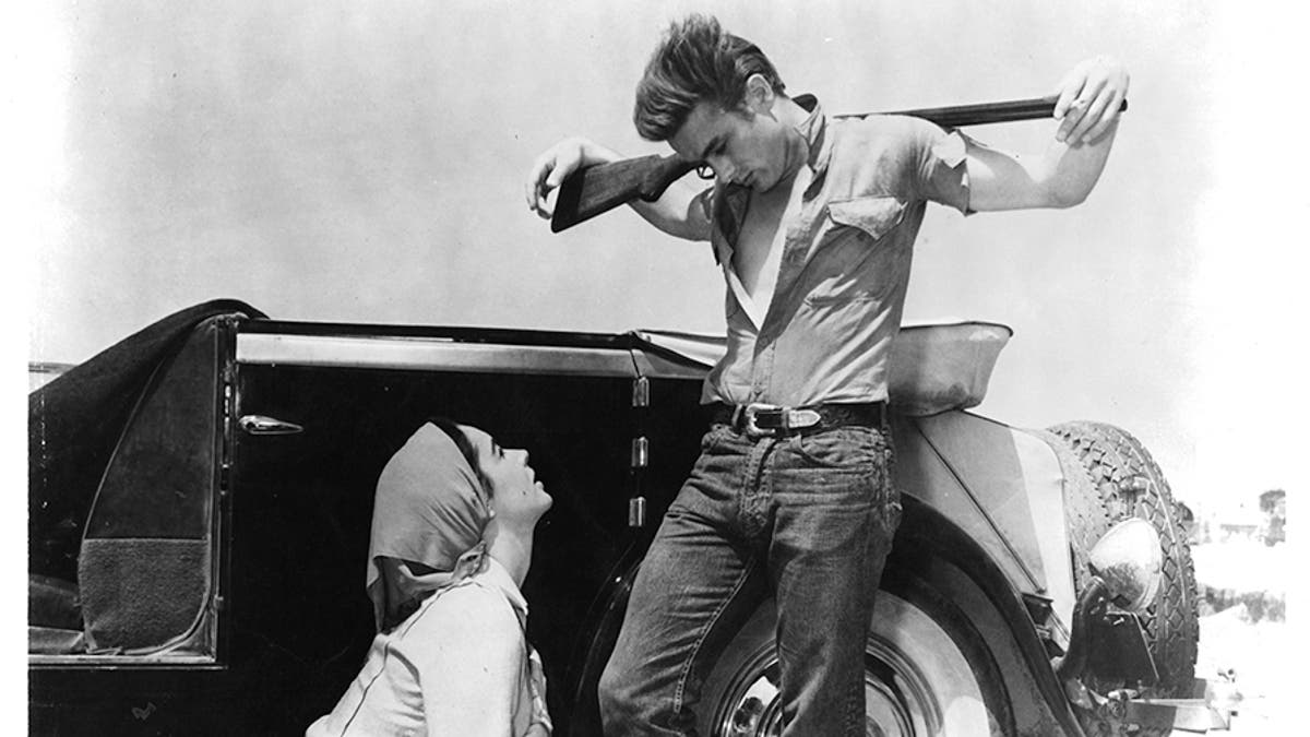 James Dean wearing an open shirt and jeans holding a shotgun and looking down at Elizabeth Taylor