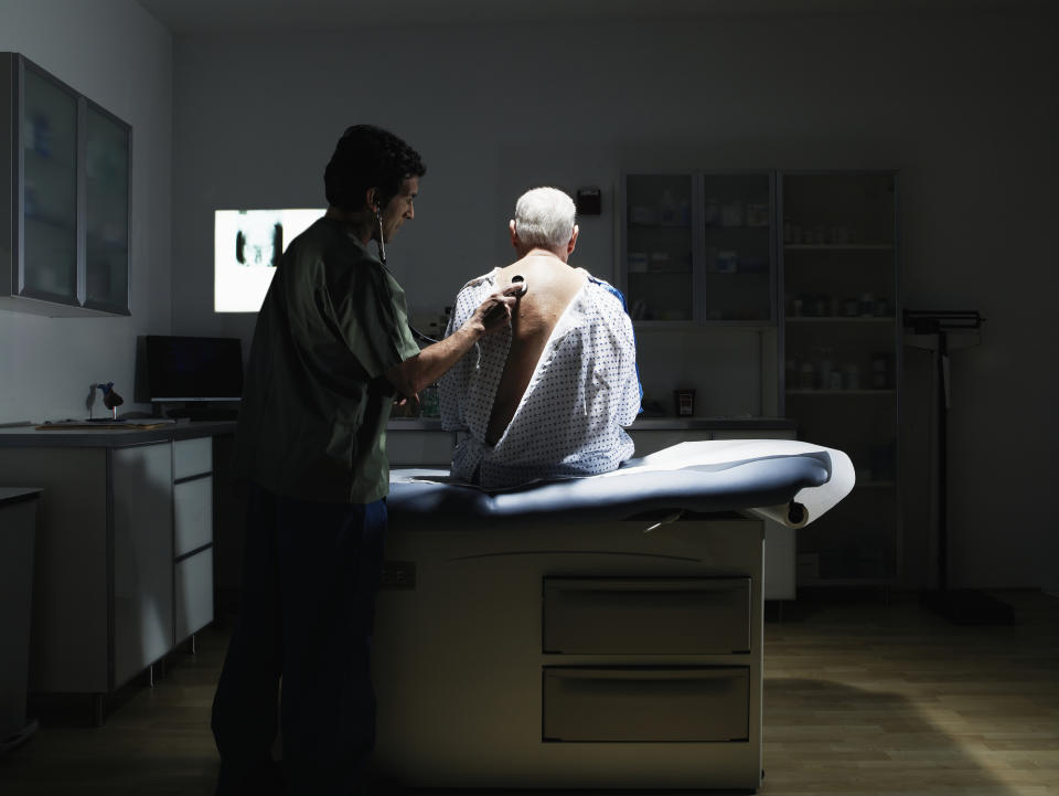 A man in a doctor's appt gown sitting on an exam table while the doctor listens to his heart with a stethoscope