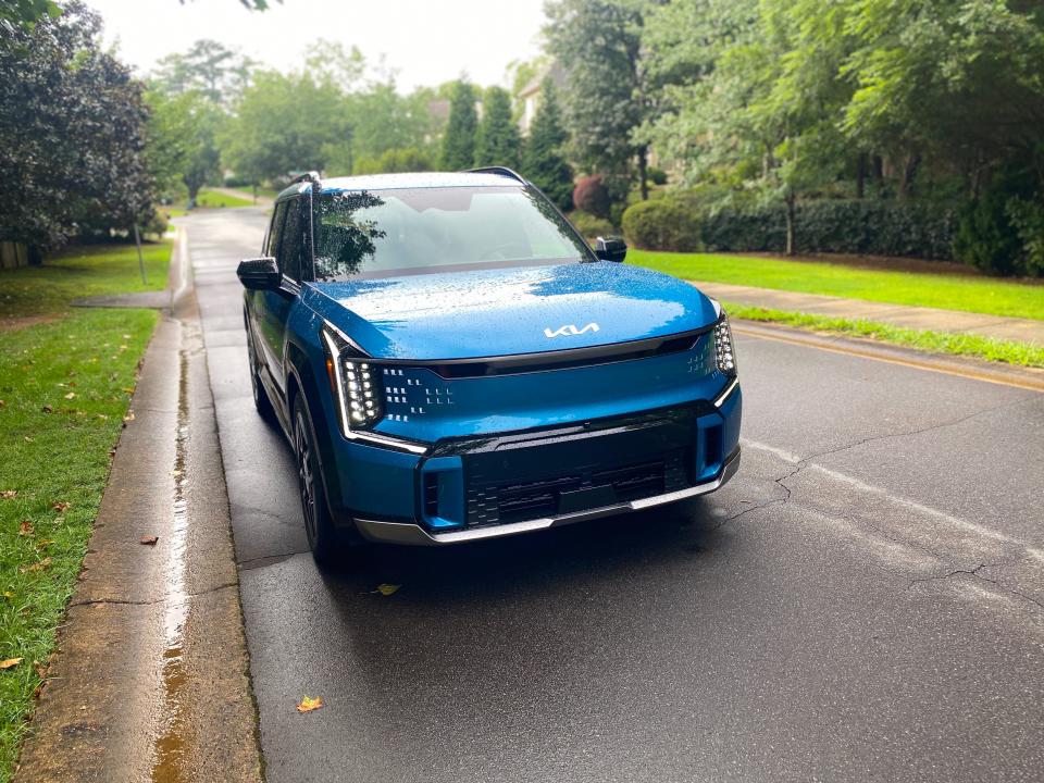 A blue 2024 Kia EV9 GT-Line EV SUV from the left front side while parked on the side of a tree-lined street.