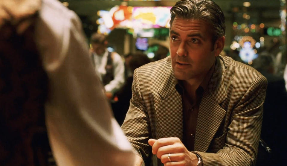 Man in a suit sits at a poker table, engaging in conversation with another person, in a lively casino setting