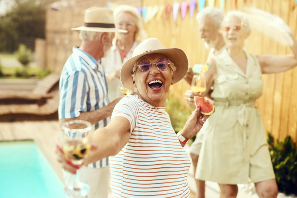 Elderly woman with a wide smile enjoying a party outdoors with friends, wearing a hat and sunglasses