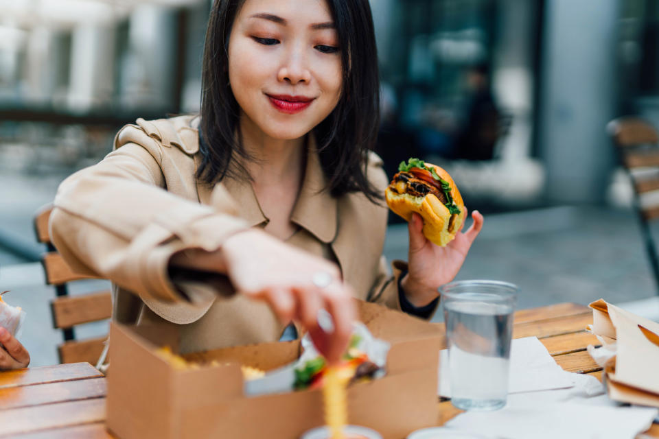 A woman enjoys a burger outside at a wooden table, wearing a trench coat, with a to-go food box and a beverage next to her