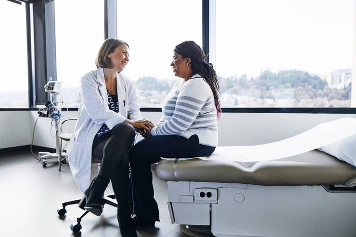 A doctor and patient sit smiling in a well-lit medical office. The doctor, in a white coat, listens attentively. The patient wears a striped sweater