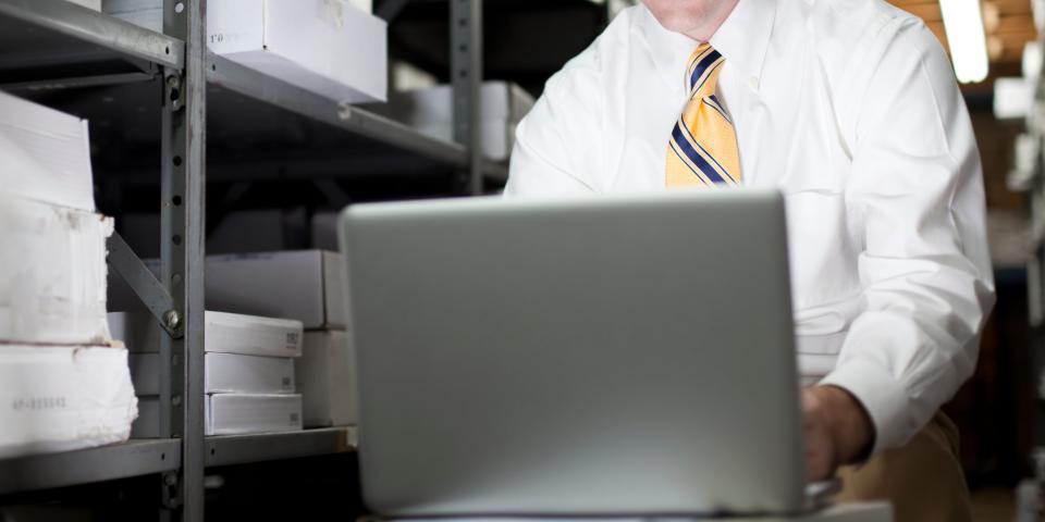 Man in a white shirt with a yellow tie works on a laptop in a storage room with shelves of boxes