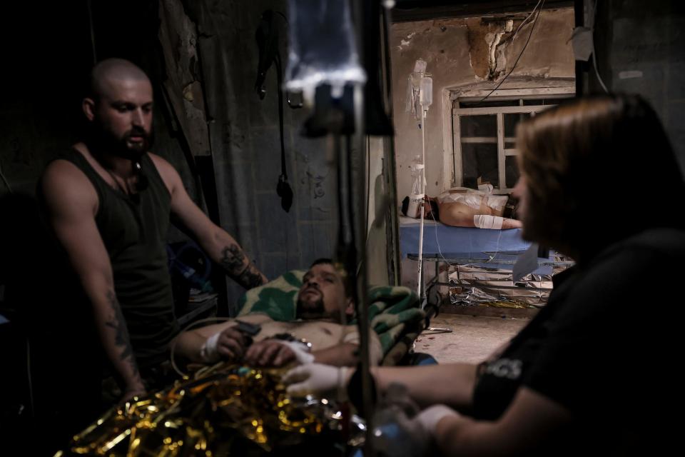 Two medics stand over a Ukrainian soldier on a bed with an IV in a dark room with exposed brick