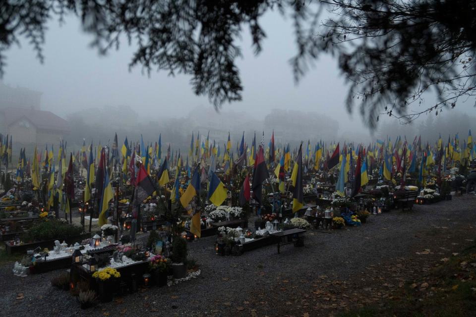 Graves are seen in a misty graveyard, with Ukrainian flags, candles, and flowers on them.