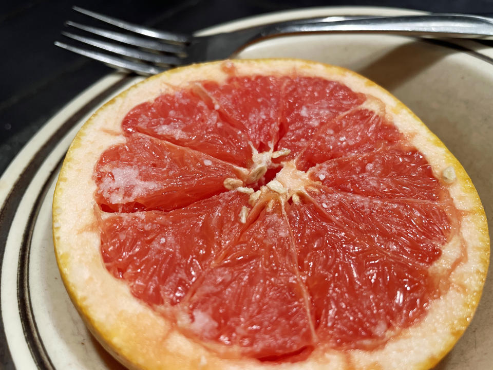 A halved grapefruit, showing the juicy sections and some seeds inside, sits on a plate with a fork next to it