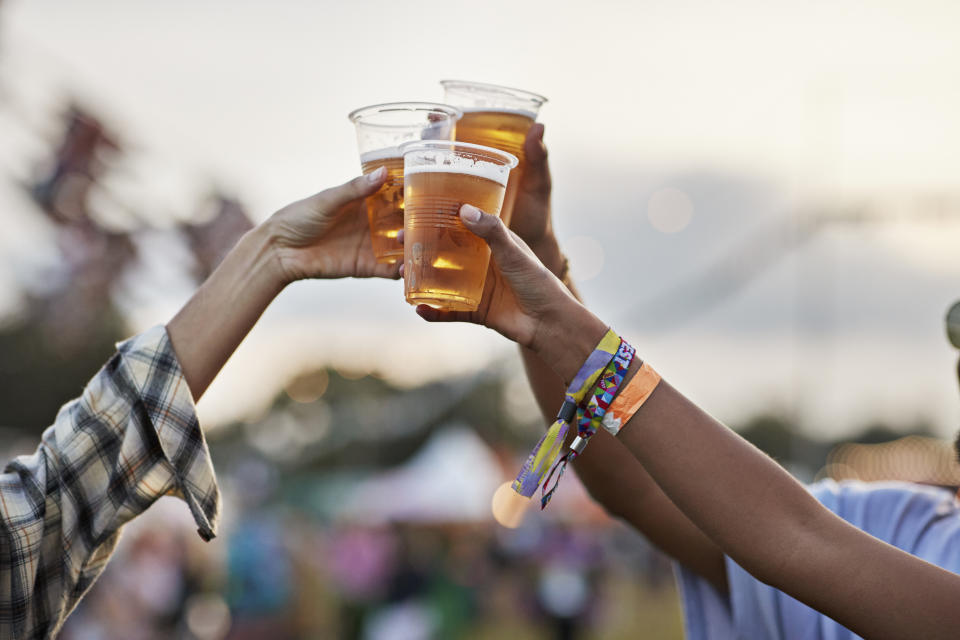 People clinking plastic cups of beer outdoors at a festival, wearing wristbands, with a blurred background of tents and lights