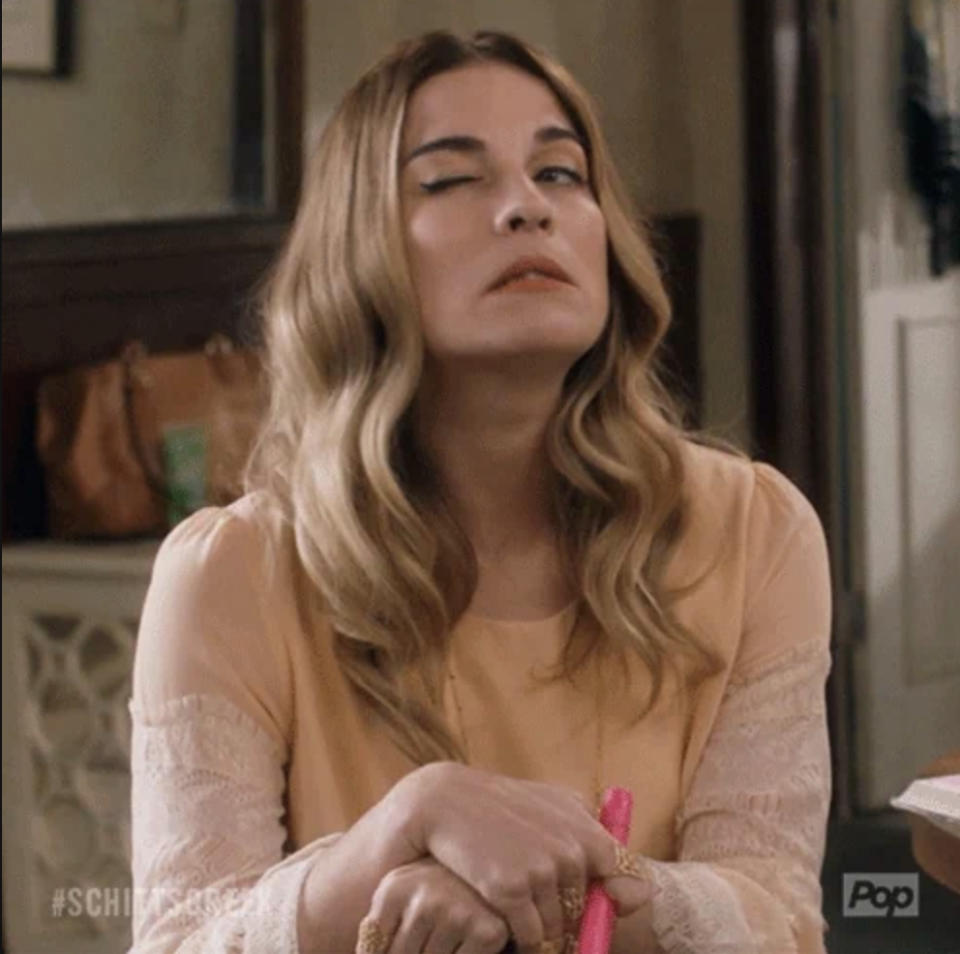 Person with long hair winks and holds a pink object, sitting indoors. Text &quot;SCHITT'S CREEK&quot; appears in the corner