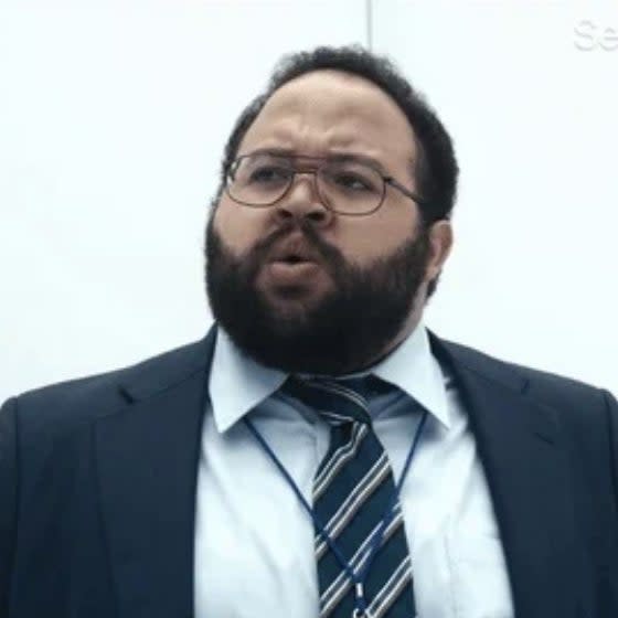 Person with a beard and glasses in a suit and striped tie, appearing surprised or concerned, with a plain background