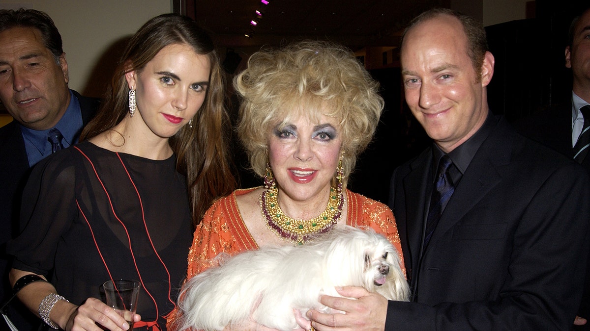 Elizabeth Taylor in between a woman and a man in black as she wears an orange dress and holds a small white dog