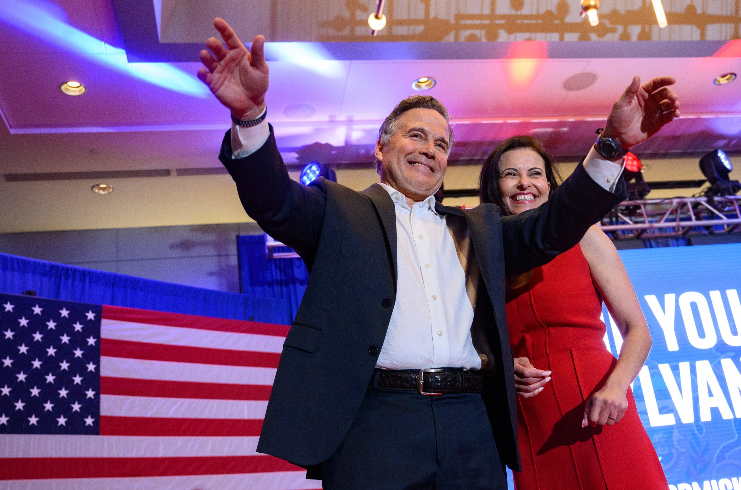 A politician and his wife smile and gesture toward a crowd, with a US flag behind them.