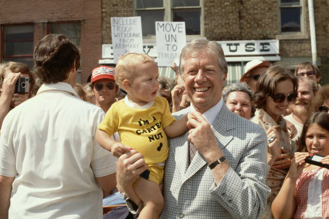 President Carter poses with a baby in Quincy, Ill., in 1979, during a summer vacation cruising the Mississippi River. The baby's T-shirt reads "I'm a Carter Nut" above the outline of a peanut.
