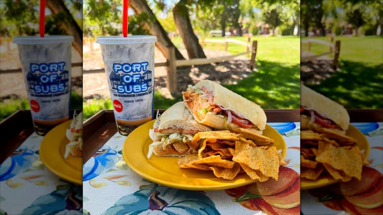 Port of Subs subway sandwich, with chips and a drink, sitting on an outside table with a sunny park in the background