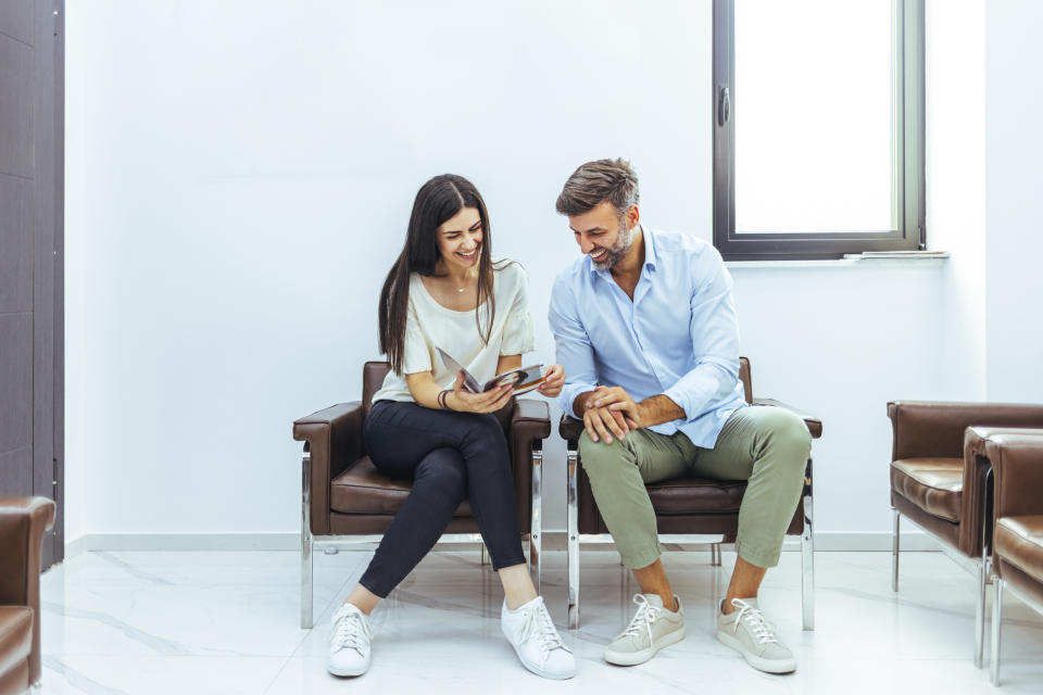 A woman and a man sitting on chairs, smiling and looking at a brochure in a bright, modern room with a large window