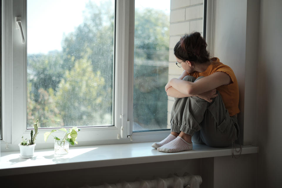 Person sitting on a windowsill, looking outside, wearing casual clothing. Indoor plants are nearby