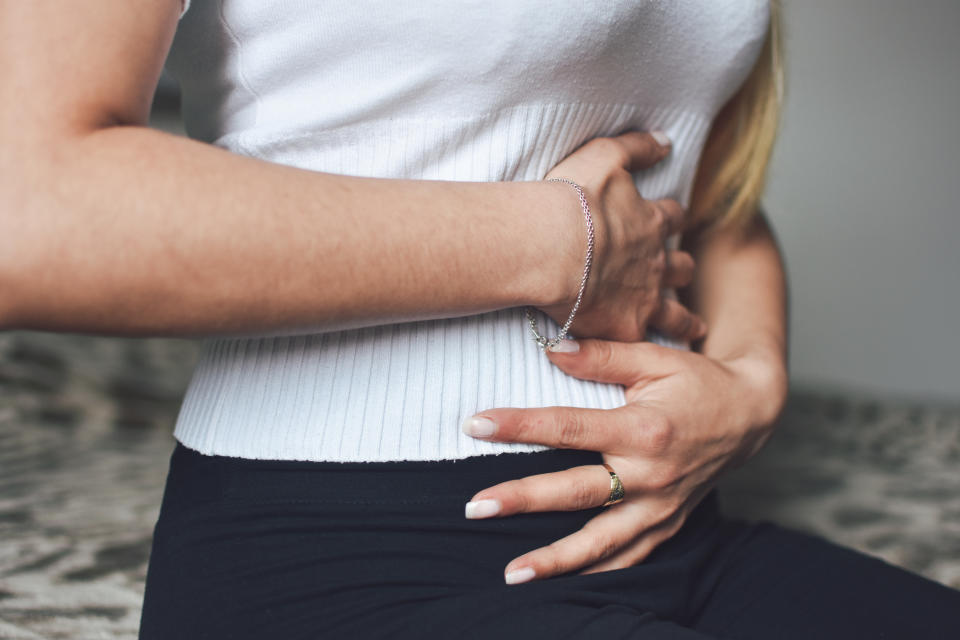 Person holding their stomach with both hands, wearing a ribbed top and bracelet, suggesting discomfort or abdominal pain