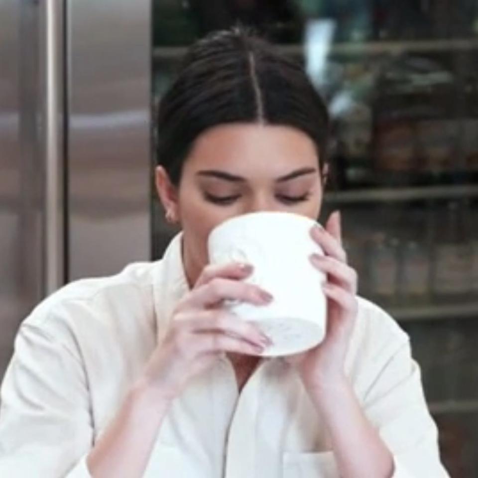 Person drinking from a white cup in a kitchen, wearing a casual white shirt