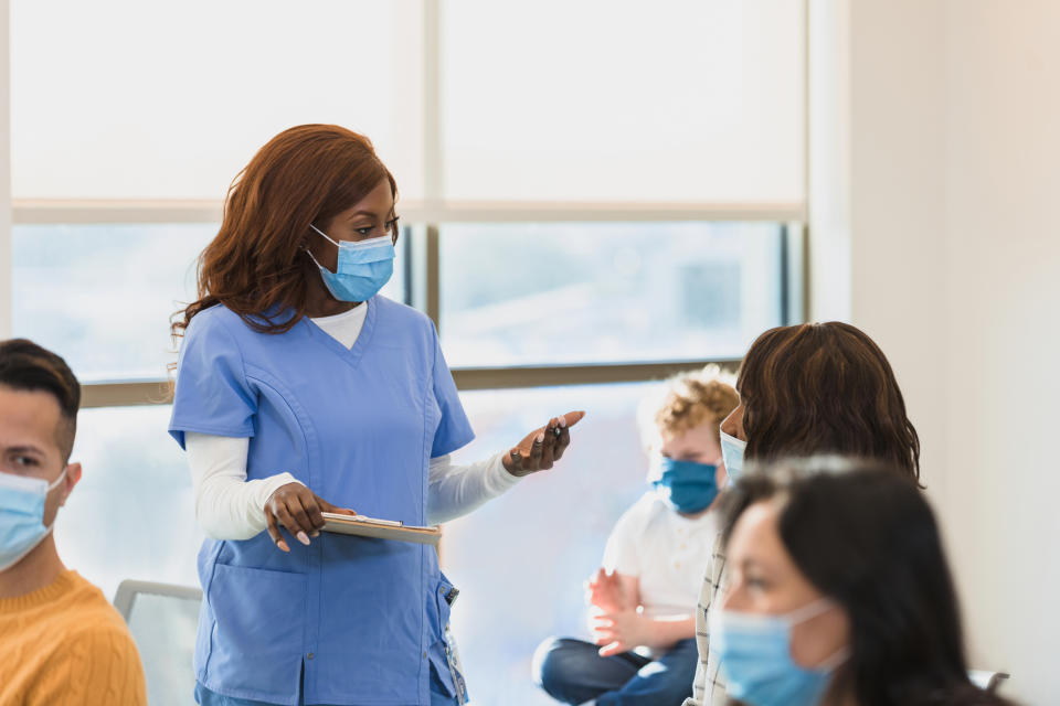 Healthcare worker in scrubs and mask speaks with a seated person in a clinic. Other masked individuals are in the background