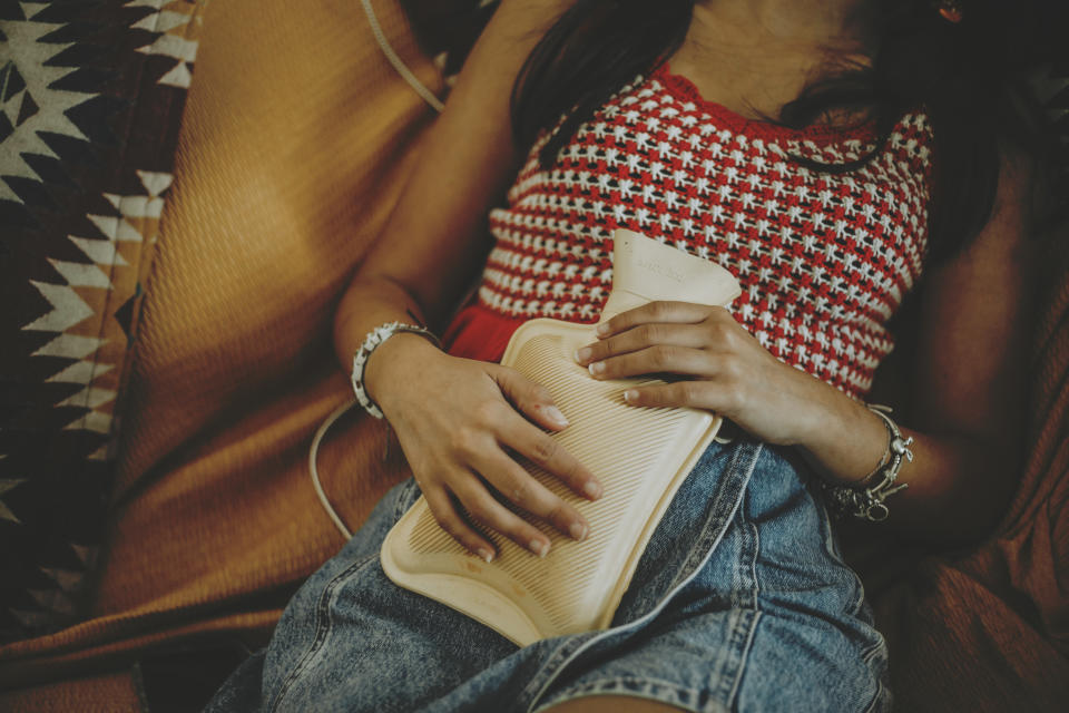 Person relaxing on a couch, wearing a patterned top and denim skirt, holding a hot water bottle on their stomach