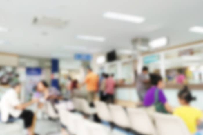 Blurry image of a busy waiting area in a hospital or clinic with people sitting on chairs and standing at the counter