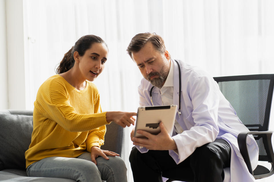 A woman consults with a doctor on a couch, both looking at a tablet, engaged in discussion