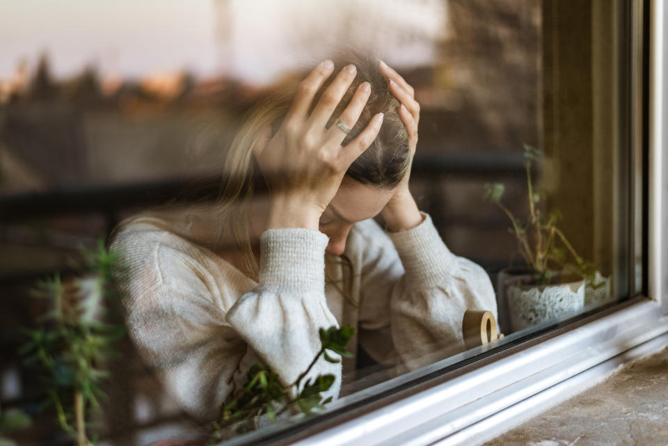 A person sits by a window with their head in their hands, appearing stressed or deep in thought. Houseplants are visible on the windowsill