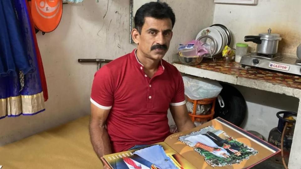 Nimisha&#39;s husband Tony Thomas wearing a red polo t-shirt and holding their wedding album is sitting on a plank placed near their kitchen. 