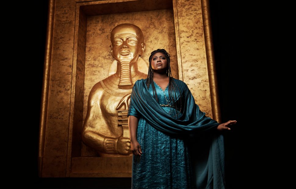 A woman in a flowing teal dress with braided hair stands confidently in front of a large golden statue of an ancient Egyptian figure, framed by a gilded backdrop, evoking the grandeur and themes of Verdi's Aida.