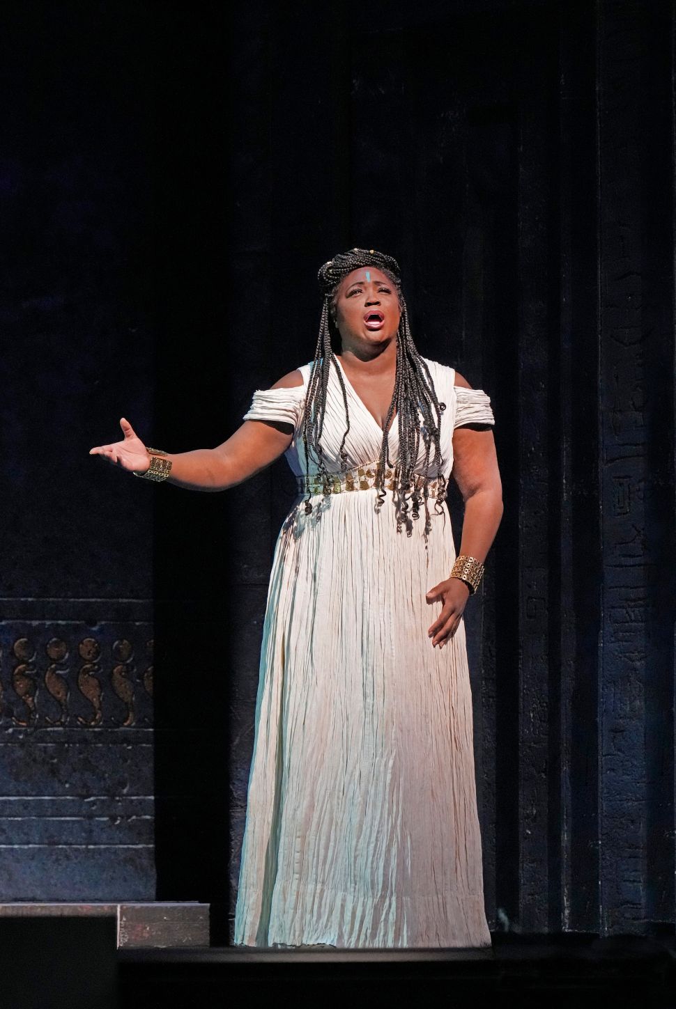 A woman dressed in a white, pleated gown with gold accessories performs on stage, her hand outstretched as she sings passionately against a dark, dramatic backdrop with Egyptian-inspired motifs.