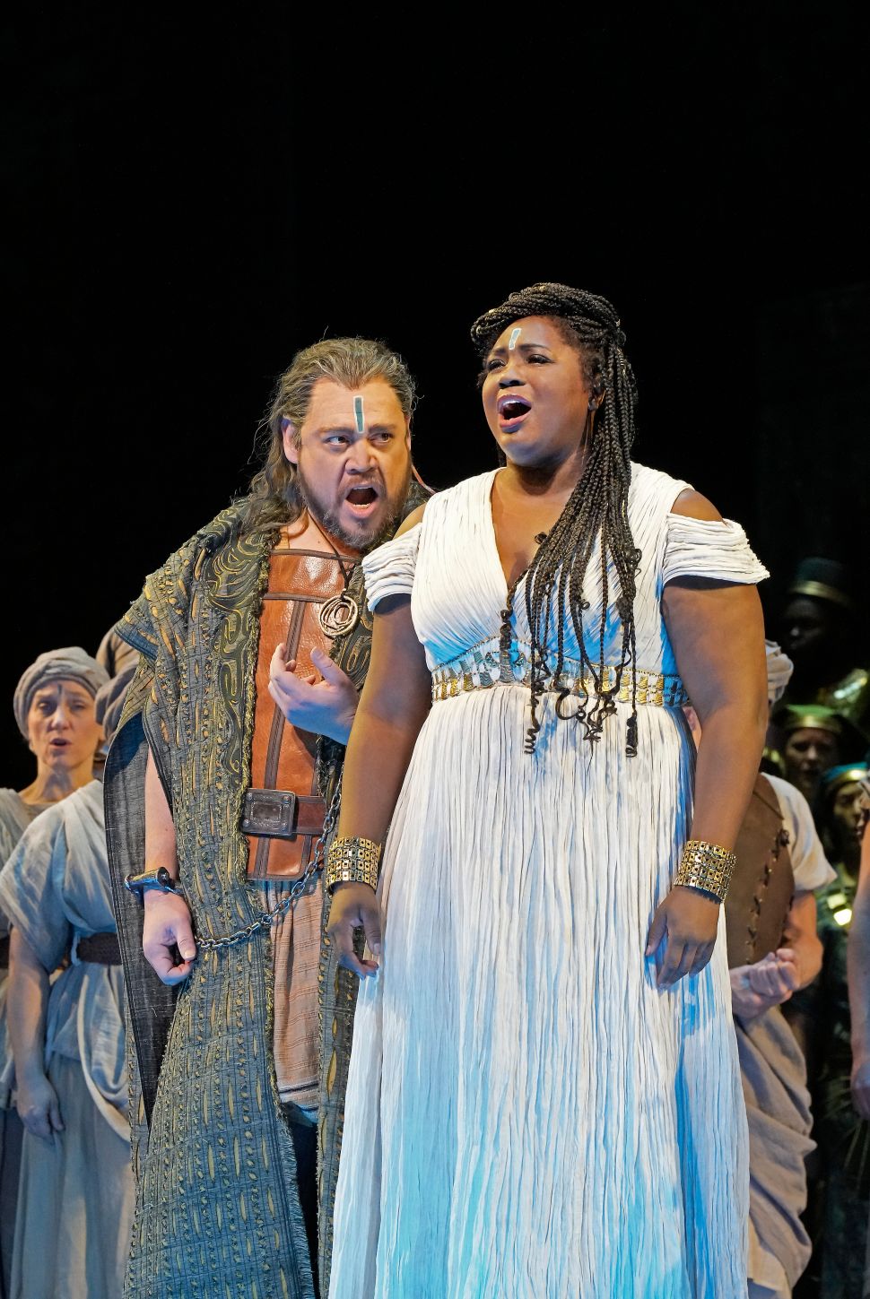 A man in a detailed costume stands beside a woman in a white gown with braided hair, both singing with intense expressions during a theatrical performance, surrounded by an ensemble cast in historical costumes.