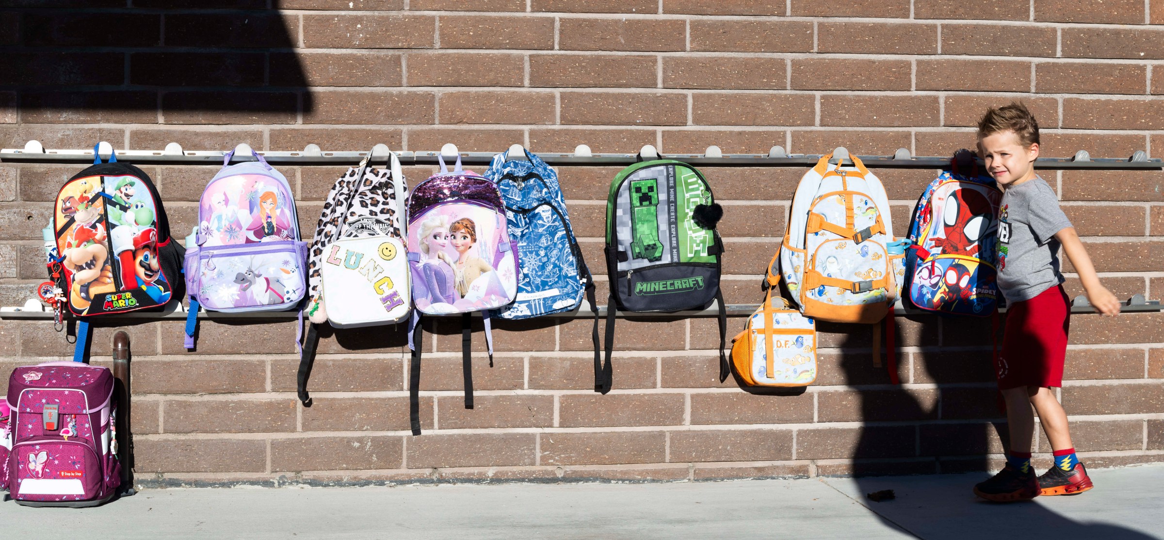 A young boy places a Spiderman backpack on a rack on a brick wall alongside other backpacks