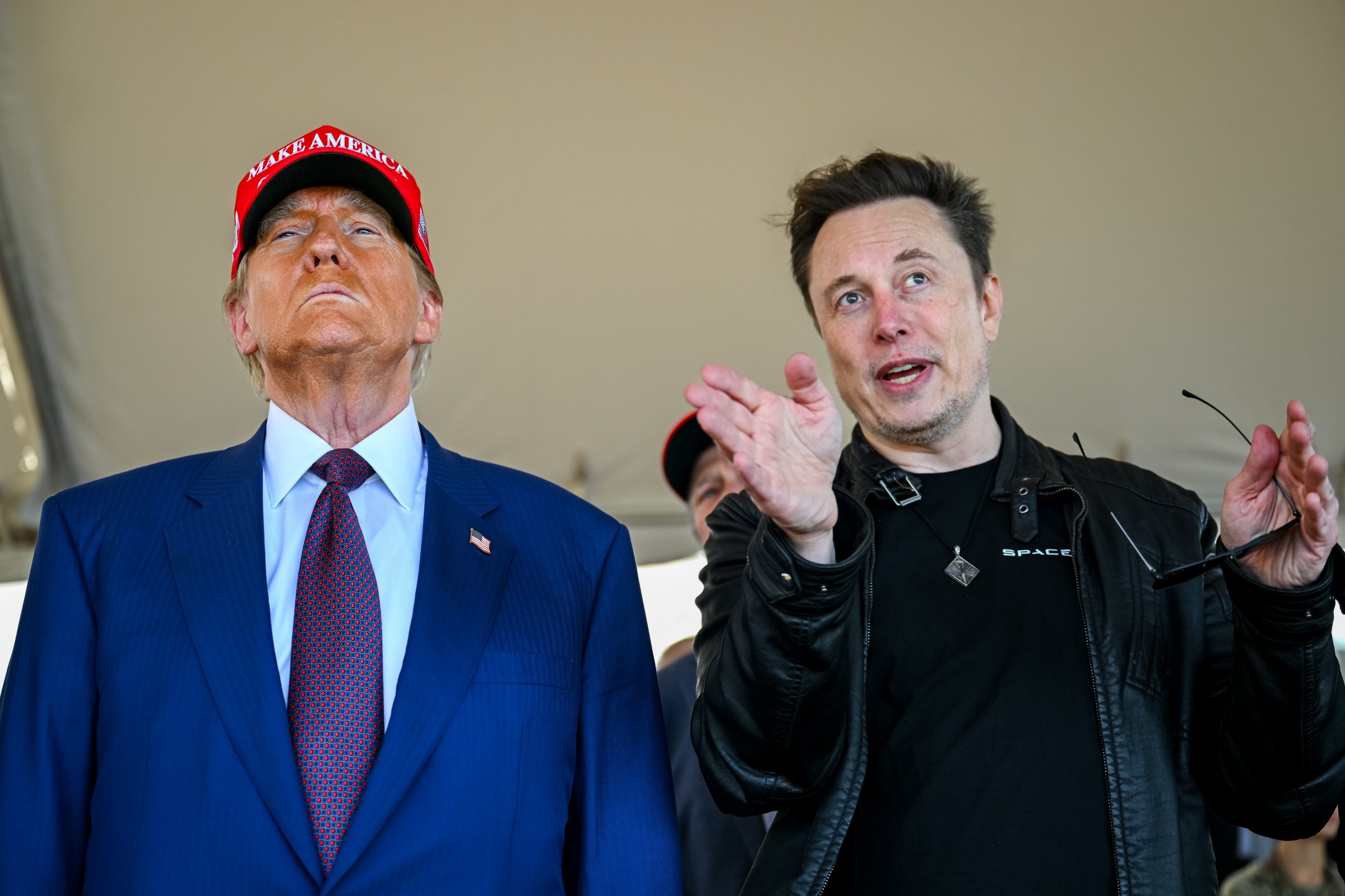 Trump looking ahead in a MAGA hat while Elon talks and gestures beside him.