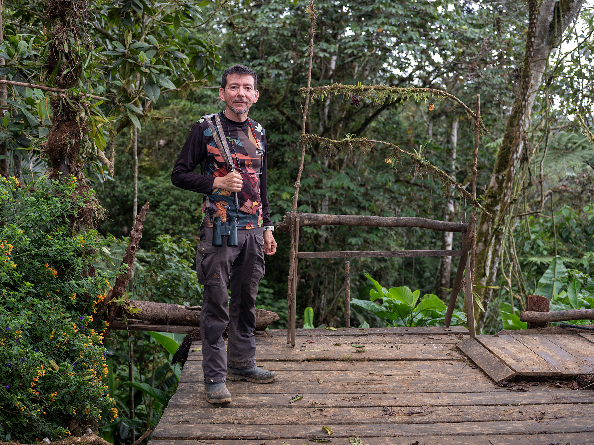 A man holding binoculars stands on a wooden platform with greenery surrounding