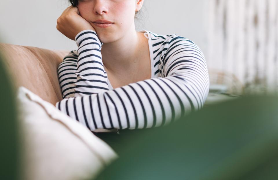 Person with a thoughtful expression sits on a couch. They are wearing a striped shirt and resting their head on their hand