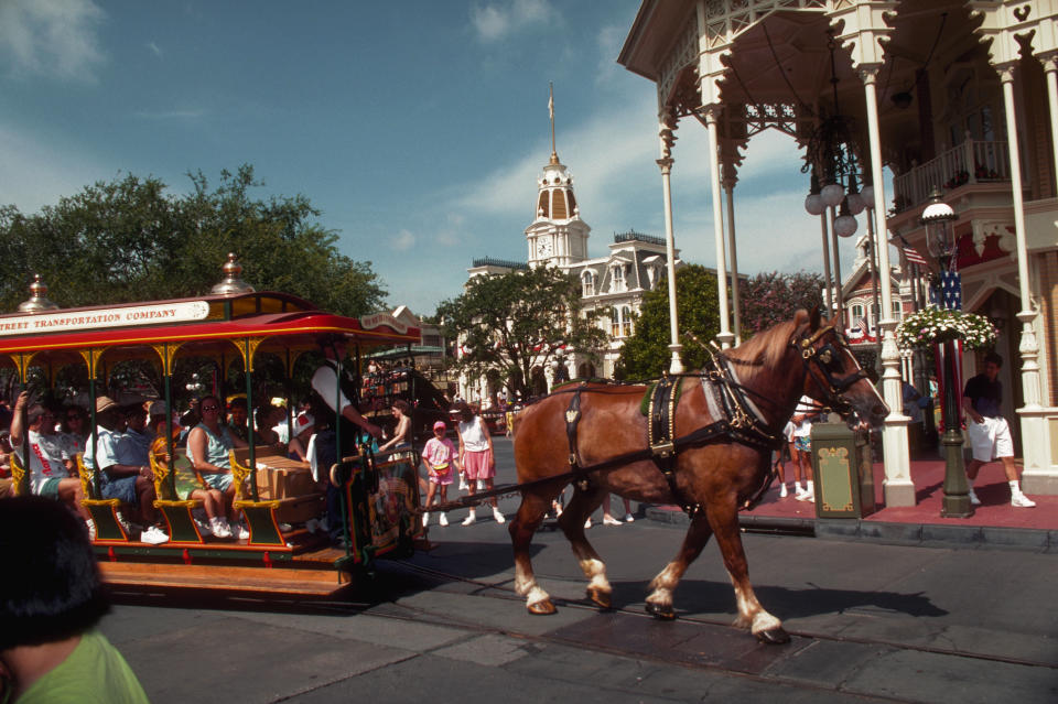 Horse pulling a trolley with passengers through a bustling street scene, featuring historic buildings and an outdoor gathering of people